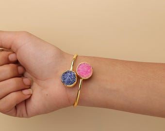 May include: A gold-tone open cuff bracelet featuring two circular druzy stones, one in a deep blue and the other in a vibrant pink. The stones are set in gold-tone bezels, creating a contrasting look on the thin, polished metal band.