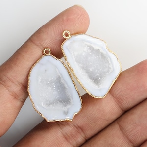 May include: Two irregular white agate geode pendants with sparkling crystalline centers and gold-toned metallic edges. Each pendant features a small circular gold-toned jump ring at the top for attachment, showcasing a natural stone texture.