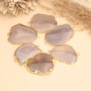 May include: A collection of five irregular, smooth, light gray agate stone slices, each featuring a polished surface and a metallic gold-toned electroplated edge. Every stone slice includes two small gold-toned metal loops attached to opposite sides.