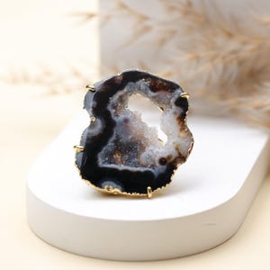 May include: A geode ring with a gold-coloured band. The ring features a polished, irregularly shaped stone with a mix of black, white, and brown colours. The stone has a hollow centre with crystal formations.