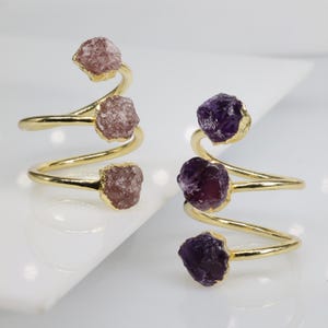Op de afbeelding: Twee gouden ringen met drie ruwe edelsteen accenten. De ene ring heeft roze stenen en de andere heeft paarse stenen.