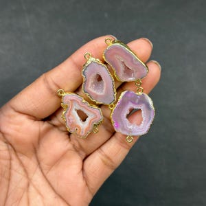 Puede incluir: Cuatro colgantes de piedra de ágata rosa y morada con bordes dorados. Cada colgante tiene una forma única y un pequeño agujero en la parte superior para ensartar. Los colgantes se muestran sobre un fondo oscuro.
