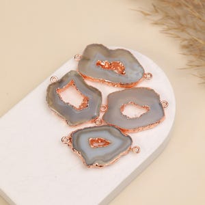 May include: Four irregular gray agate slices with rose gold electroplated edges. Each stone features a central geode cavity with sparkling crystals and two small metal loops attached to the sides for use as jewelry connectors.