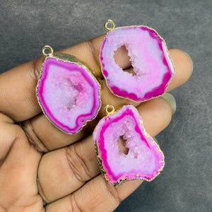 May include: Three pink and white agate pendants with gold-colored edges and loops. The pendants have a geode-like appearance with a hollow center. The pink color is vibrant, and the gold edges add a touch of elegance. These pendants are ideal for jewelry making.