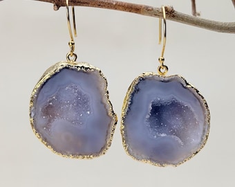 Puede incluir: Un par de pendientes colgantes con rodajas circulares irregulares de piedra ágata de color morado claro. Cada piedra tiene un centro cristalino texturizado y está enmarcada por un borde fino de color dorado. Cuelgan de ganchos dorados.