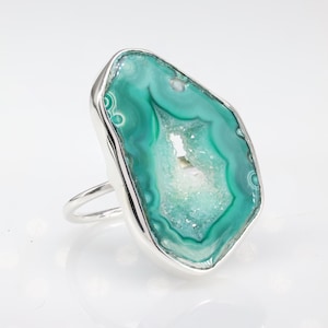 May include: A silver ring featuring a large, green agate stone. The agate has a natural, crystal-like formation with a hollow centre.