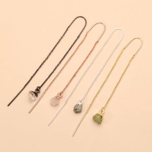 Peut inclure: Quatre boucles d'oreilles pendantes composées de fines chaînes métalliques aux tons noir, or rose, argent et or. Chaque boucle est terminée par une petite pierre brute de forme irrégulière fixée à l'extrémité de la chaîne, révélant des textures naturelles.