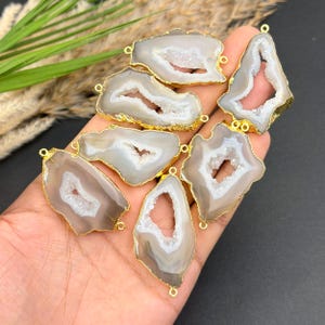 May include: A collection of seven irregular, translucent gray agate slices with white crystalline centers. Each stone features a metallic gold-toned electroplated edge and two small circular loops at opposite ends for use as jewelry connectors.