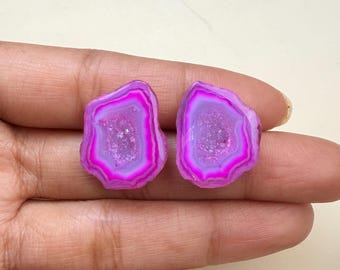 May include: Two irregular, flat-cut agate slices featuring vibrant concentric bands of hot pink and soft lavender. The centre of each stone displays a textured, crystalline druzy surface with a sparkling, light-reflecting finish and jagged edges.