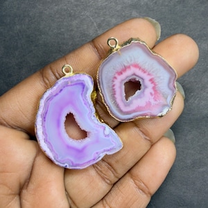 May include: Two natural agate stone pendants with gold-colored edges. One pendant is lavender and white, the other is pink and white. Both have a hole in the center and a small loop at the top for hanging.