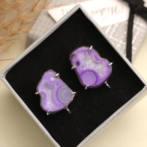 May include: A pair of purple agate earrings with silver-colored prongs, presented in a black velvet-lined gift box. The earrings have a natural, organic shape with varying shades of purple and white.