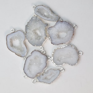 Pode incluir: Uma coleção de fatias de ágata brancas e cinzentas com bordas de metal prateado. Cada fatia tem uma forma única e um pequeno laço para prender a joias ou outros trabalhos manuais. A pedra natural tem um centro cristalino.