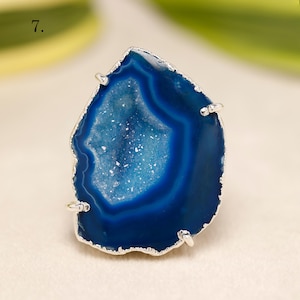 以下が含まれることがあります： シルバーカラーの金属セッティングが施された、大きな形の不規則な青い瑪瑙の指輪。瑪瑙は、きらめく結晶質の内部を持つジオードの中心部を備えています。リングのデザインは、石の自然な美しさを際立たせています。