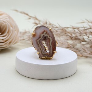 May include: A gold-toned ring featuring a large, polished agate stone. The agate displays a natural pattern of brown, white, and cream hues, with a central cavity. The ring is set on a white pedestal, with dried plants in the background.