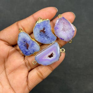May include: Four polished, natural agate pendants in shades of blue and purple, with gold-colored metal edging and small loops for jewelry making. The pendants have a geode-like appearance with a hollow center.