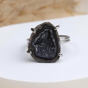Peut inclure: Bague en argent ornée d'une pierre géode gris foncé, avec un centre scintillant rempli de cristaux. La pierre en forme de goutte d'eau est maintenue par quatre griffes en argent. La bague a un design simple à double anneau.