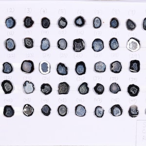 May include: Fifty small, polished stones with dark edges and lighter, crystalline centres, arranged in a grid. Each stone is an irregular shape. The size is 1.5 cm. The stones are displayed on a white surface.
