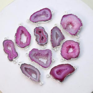 Puede incluir: Una colección de colgantes de piedras preciosas de ágata rosa y púrpura con bordes y bucles de metal plateado. Los colgantes varían en forma y tamaño, mostrando los patrones y colores naturales de las piedras. Ideal para la creación de joyas.