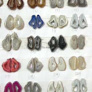 May include: Assortment of polished agate stone pairs in various colors, including brown, blue, gray, and pink. Each stone is cut in half, revealing a crystal-lined center. The stones are numbered for inventory.