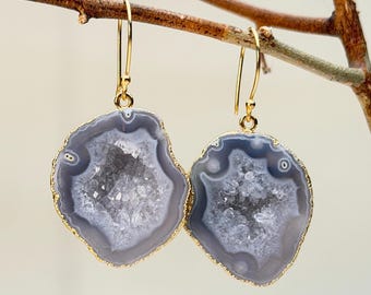 Puede incluir: Un par de pendientes colgantes dorados con rodajas de ágata pulida gris y blanca y un borde dorado. Los pendientes cuelgan de una rama marrón sobre un fondo suave y neutro. Joyería natural única.