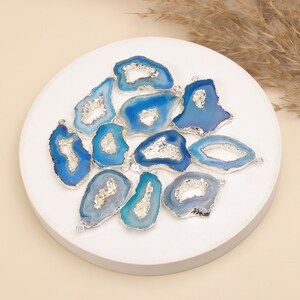 May include: A collection of irregular blue agate stone pendants with silver-toned metallic edges. Each piece features a central geode-like crystalline cavity and a small metal loop at the top for attachment, showcasing various shades of blue and white.