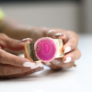 May include: A gold-tone cuff bracelet featuring a large, vibrant pink agate stone. The agate has concentric bands of pink and white. The cuff has a textured surface. A stylish piece of jewelry.