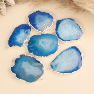 May include: A collection of five irregular, flat blue agate stone slices with silver-toned metal edges. Each stone features a translucent, crystalline center and a deep blue outer rim, with small metal loops attached to the sides for jewelry making.