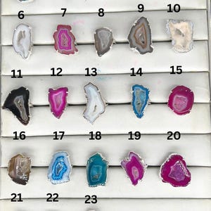 May include: Twenty-three agate rings displayed in a numbered grid. The rings are made of silver and feature a variety of colors, including blue, pink, white, and black.