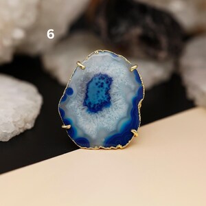 May include: A large, natural agate ring with a gold-colored band. The agate features concentric bands of white, light blue, and deep blue. The number "6" is visible in the upper left corner.