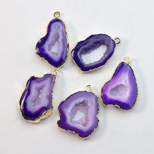 May include: Five purple and white agate pendants with gold-colored edges and small loops for hanging. The pendants have a natural, geode-like appearance, with a polished finish. The color palette includes shades of purple, white, and gold.