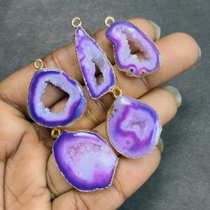 May include: Six purple and white agate pendants with gold-colored edges and loops. The pendants vary in shape, including oval, teardrop, and irregular forms. The agate has a crystalline center and a polished, smooth surface.