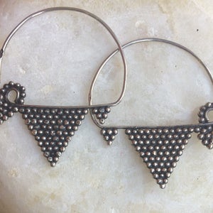 Può includere: Un paio di orecchini a cerchio d'argento con un design triangolare. Gli orecchini sono realizzati con piccole perline d'argento disposte a forma di triangolo. Gli orecchini sono appesi a un cerchio d'argento.