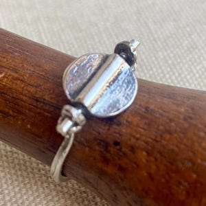 Può includere: Un anello d'argento dal design unico. L'anello presenta un elemento centrale circolare e appiattito con una linea verticale al centro. La fascia ha un dettaglio a forma di nodo su ciascun lato dell'elemento centrale. L'anello è esposto su una superficie di legno.