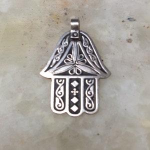 Può includere: Ciondolo a forma di mano di Hamsa color argento con intricati riccioli e disegni geometrici. Il ciondolo presenta un anello per una catena ed è probabilmente realizzato in metallo. Il design include forme floreali e a diamante.