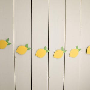 May include: A garland of yellow lemon shapes with green leaves is displayed against a white wooden backdrop. The lemons are spaced out and strung together, creating a bright, summery decoration.