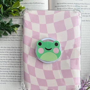 Peut inclure: Coque de téléphone transparente avec un motif à carreaux roses et blancs et un support de téléphone en forme de grenouille verte. L'étui est posé sur un livre ouvert, avec du texte visible, entouré de verdure et de fleurs violettes.