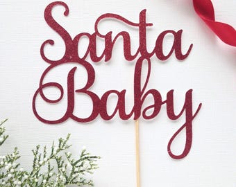 May include: A red glittery "Santa Baby" cake topper on a wooden stick. The festive decoration is set against a white background with a red ribbon and a sprig of greenery.