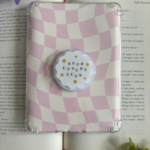 May include: A clear protective tablet case featuring a light pink and white wavy checkerboard pattern. Centered on the case is a round, scalloped-edge grip accessory with a white background, gold star accents, and the text BOOK LOVERS CLUB.