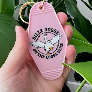 May include: A pink motel-style keyring with a gold ring. The keyring features a white goose illustration with the text "Silly Goose On The Loose Club" in a circular design. Green foliage forms the background.