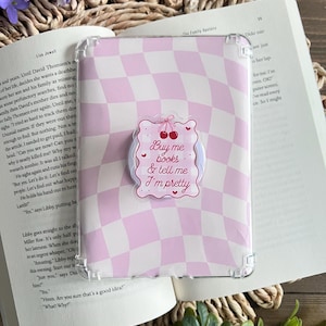 May include: A pink and white checkered phone case with a sticker that says "Buy me books & tell me I'm pretty." The case is on top of an open book with purple flowers in the background.