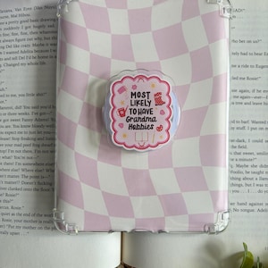 May include: A clear protective tablet case featuring a light pink and white wavy checkerboard pattern. Centered on the case is a scalloped-edge sticker with the text "MOST LIKELY TO HAVE Grandma Hobbies" surrounded by small icons of a mug, yarn, and paint.
