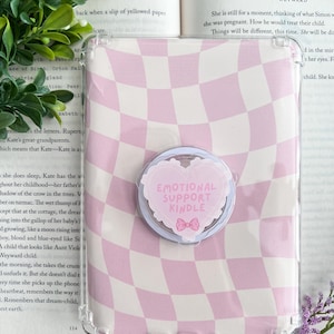May include: Transparent Kindle case featuring a pink and white chequered design. A pop socket with the text "EMOTIONAL SUPPORT KINDLE" and a pink bow is attached. The Kindle is open to a book.