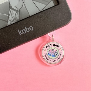 May include: A clear acrylic bookmark with a circular design, attached to a black e-reader. The bookmark features the text "PLOT TWIST: I bought more books" around a stack of books tied with a pink ribbon, set against a pink background.