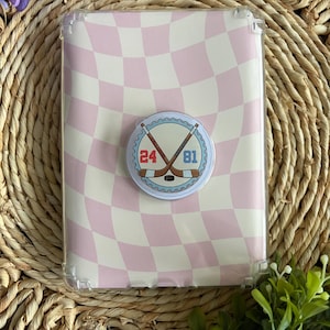May include: A clear phone case with a pink and white checkered pattern. A round sticker with crossed hockey sticks and the numbers 24 and 81 is attached to the case. The case is on a woven surface.