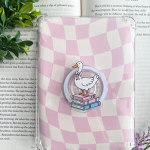May include: A transparent phone case with a pink and white chequered design. A clear pop socket shows a white goose with a bow tie, standing on books. The case rests on an open book.