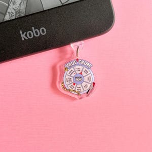 Peut inclure: Un porte-clés True Crime Mood Wheel en acrylique transparent avec un anneau argenté. Le porte-clés présente un design circulaire avec divers termes liés aux faits divers et un petit graphique de couteau rouge. Le fond est rose uni.