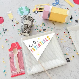 May include: A collection of school-themed decorations and supplies, including a paper cup, a pencil-shaped container, and a banner that says "hello essentials". The scene is decorated with confetti and other school-related items.