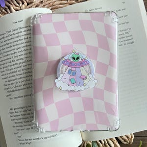 May include: Clear phone case with a pink and white checkered pattern. A decorative pop socket featuring an alien in a UFO, surrounded by clouds, books, and hearts, is attached to the case. The case is resting on an open book.