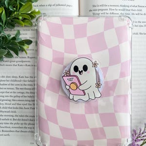 May include: Clear phone case with a pink and white checkered pattern. A cartoon ghost-shaped pop socket with the text "BOOKISH GIRLUE" is attached. The ghost is holding a book and decorated with small flowers.