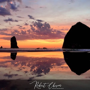 May include: Silhouette of Haystack Rock and two smaller rocks at sunset with a reflection in the water. The sky is a vibrant orange and pink with clouds.